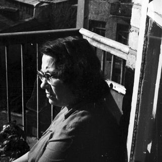 Woman in glasses stands on a balcony overlooking rooftops.