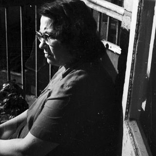 Woman in glasses stands on a balcony overlooking rooftops.