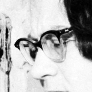 Woman with glasses sits near a brick wall and window.