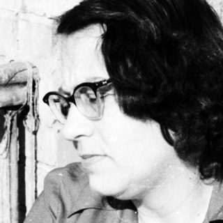 Woman with glasses sits near a brick wall and window.