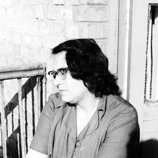 Woman with glasses sits near a brick wall and window.