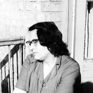 Woman with glasses sits near a brick wall and window.