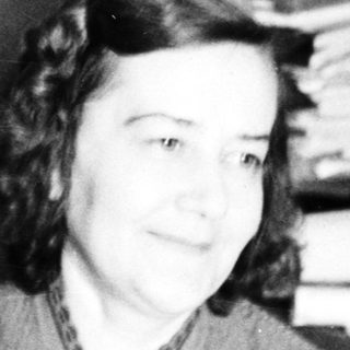 Here's a brief summary of the image: Woman seated near a desk with papers and belongings.