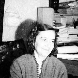 Here's a brief summary of the image: Woman seated near a desk with papers and belongings.