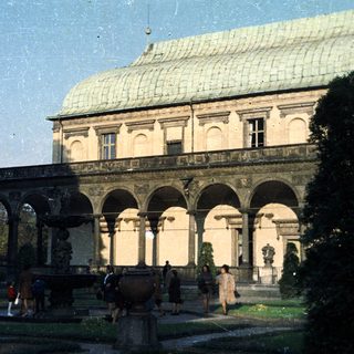 Palace facade with colonnade, gardens, and people present.