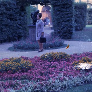 Woman in a coat stands near flowerbeds and a statue.