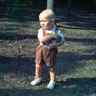 Here's a brief summary of the image: Toddler in overalls stands outdoors, holding a toy.
