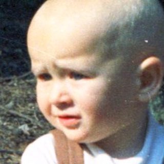 Here's a brief summary of the image: Toddler in overalls stands outdoors, holding a toy.