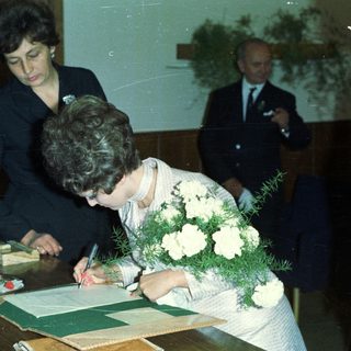 Woman signing document, likely a marriage certificate, with onlookers.