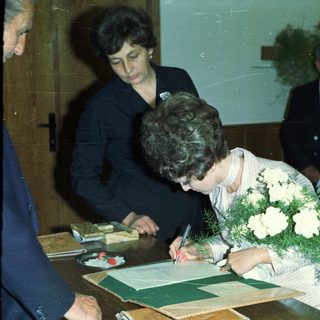 Woman signing document, likely a marriage certificate, with onlookers.