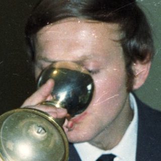 Man drinks from a horn, surrounded by people.