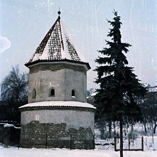 Snow-covered building with a conical roof and evergreen tree.