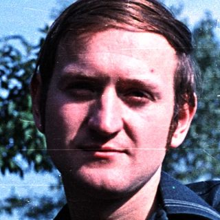 Man with short hair stands outdoors with trees in background.