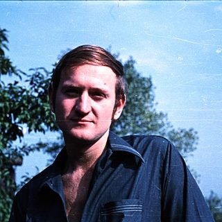 Man with short hair stands outdoors with trees in background.