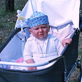 Baby in a vintage stroller sits outdoors, looking upset.