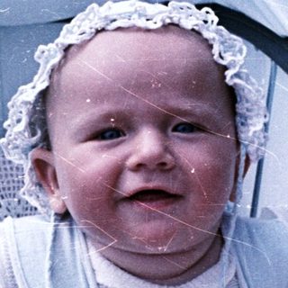 Infant sits in stroller, wearing a bonnet and outfit.