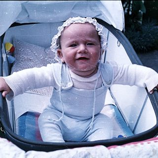 Infant sits in stroller, wearing a bonnet and outfit.