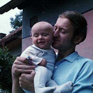 Man holds baby, smiling, in an outdoor setting.
