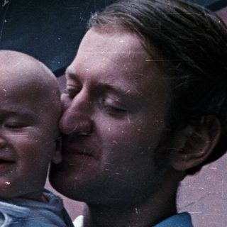 Man holds baby, smiling, in an outdoor setting.