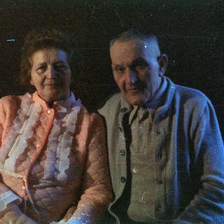 An elderly couple sits near a candlelit cabinet.