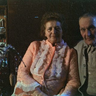 An elderly couple sits near a candlelit cabinet.