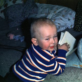 Here's a brief summary of the image: Smiling toddler sits on bed, holding a book.