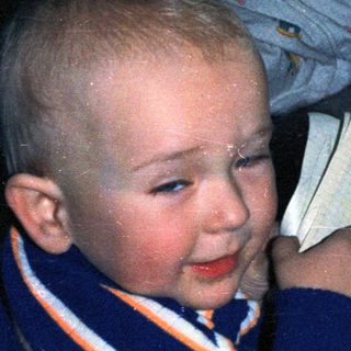 Here's a brief summary of the image: Smiling toddler sits on bed, holding a book.