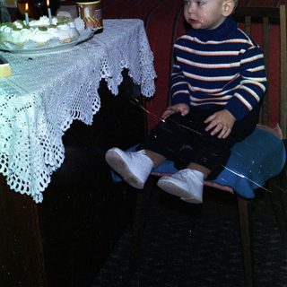 Baby sits near a table with cake and drinks.