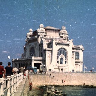 People stroll along a promenade near a grand coastal building.