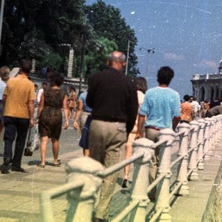People stroll along a coastal promenade with a grand building.