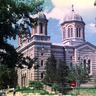 Grand stone church with domes stands amidst trees and greenery.