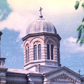Grand stone church with domes stands amidst trees and greenery.