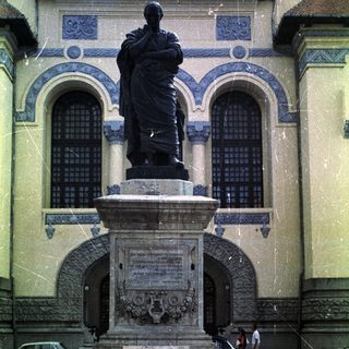 Statue on pedestal before building, with car and people visible.