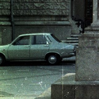 Statue on pedestal before building, with car and people visible.