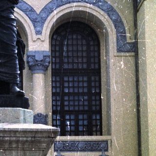 Statue on pedestal before building, with car and people visible.