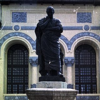 Statue on pedestal before building, with car and people visible.