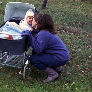 Woman crouches near a baby in a stroller outdoors.