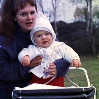 Woman with baby in stroller stands in grassy area.