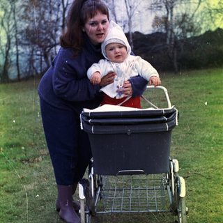 Woman with baby in stroller stands in grassy area.