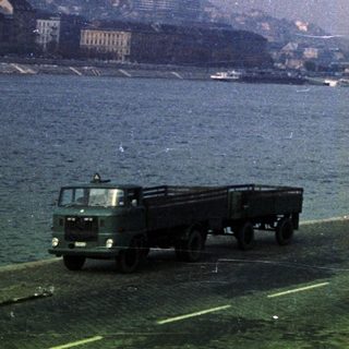 Truck drives along a river with a cityscape backdrop.
