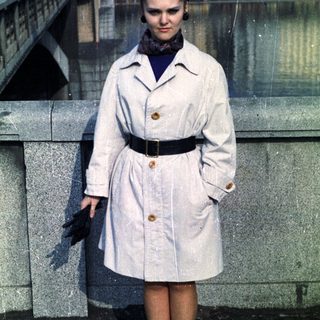 Woman in trench coat stands on bridge, looking down.