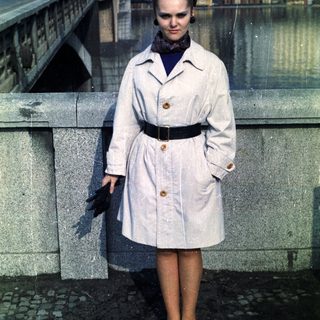 Woman in trench coat stands on bridge, looking down.