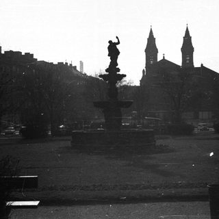 Fountain in a park with buildings and church spires.