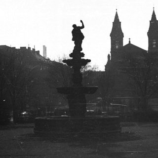 Fountain in a park with buildings and church spires.