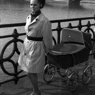 Woman stands near a pram with a river in background.