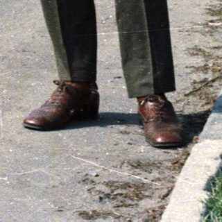 Man stands on a road near a building, trees visible.