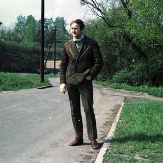 Man stands on a road near a building, trees visible.