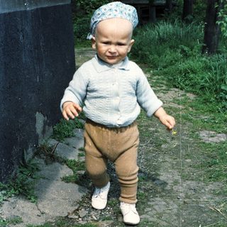 Baby in blue sweater and corduroys stands outdoors.
