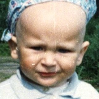 Baby in blue sweater and corduroys stands outdoors.