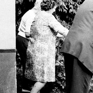Elderly couple walks outdoors, supported by a cane.